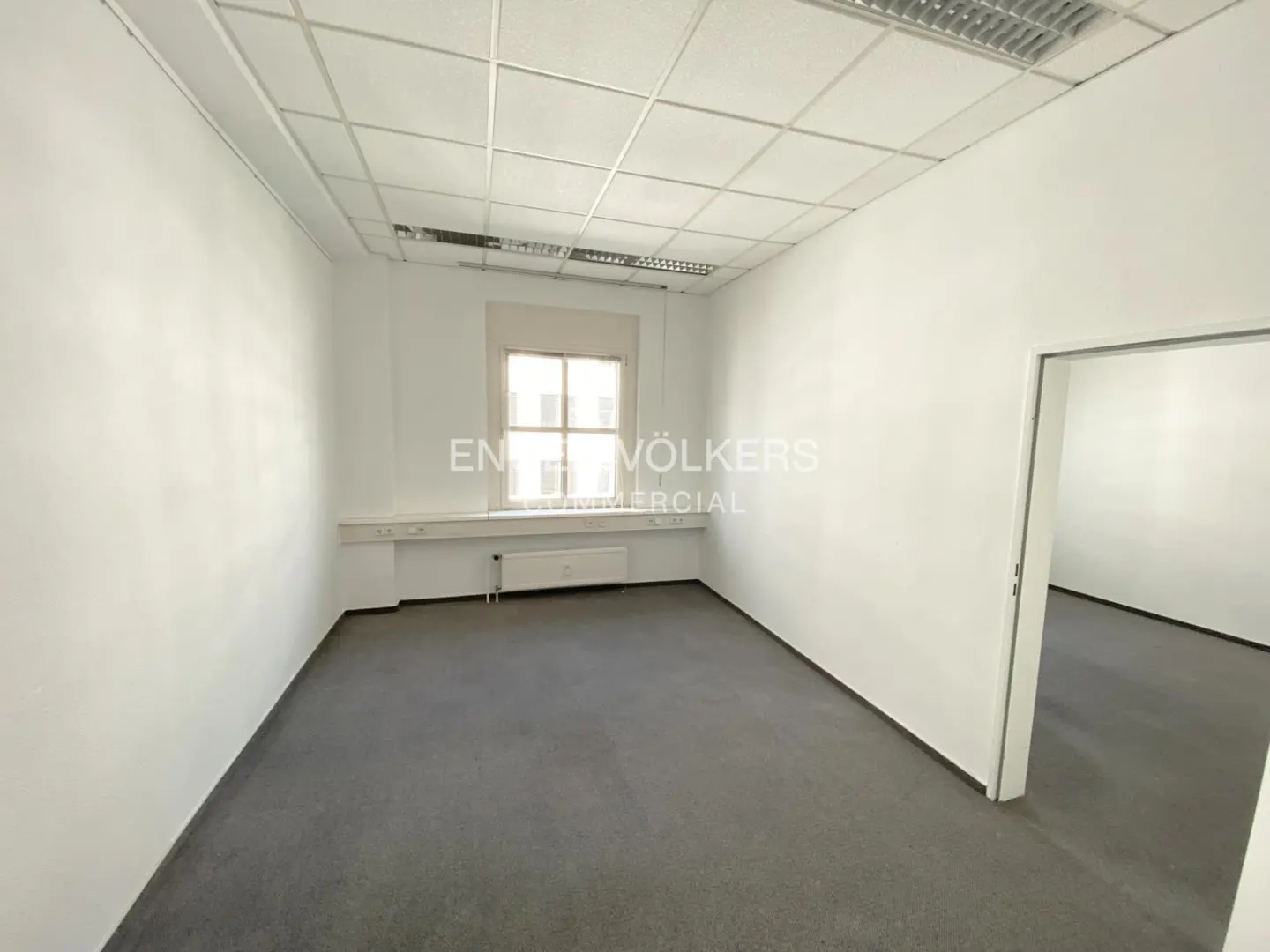 Empty office space with gray carpet, white walls, and a window. A doorway leads to another room.