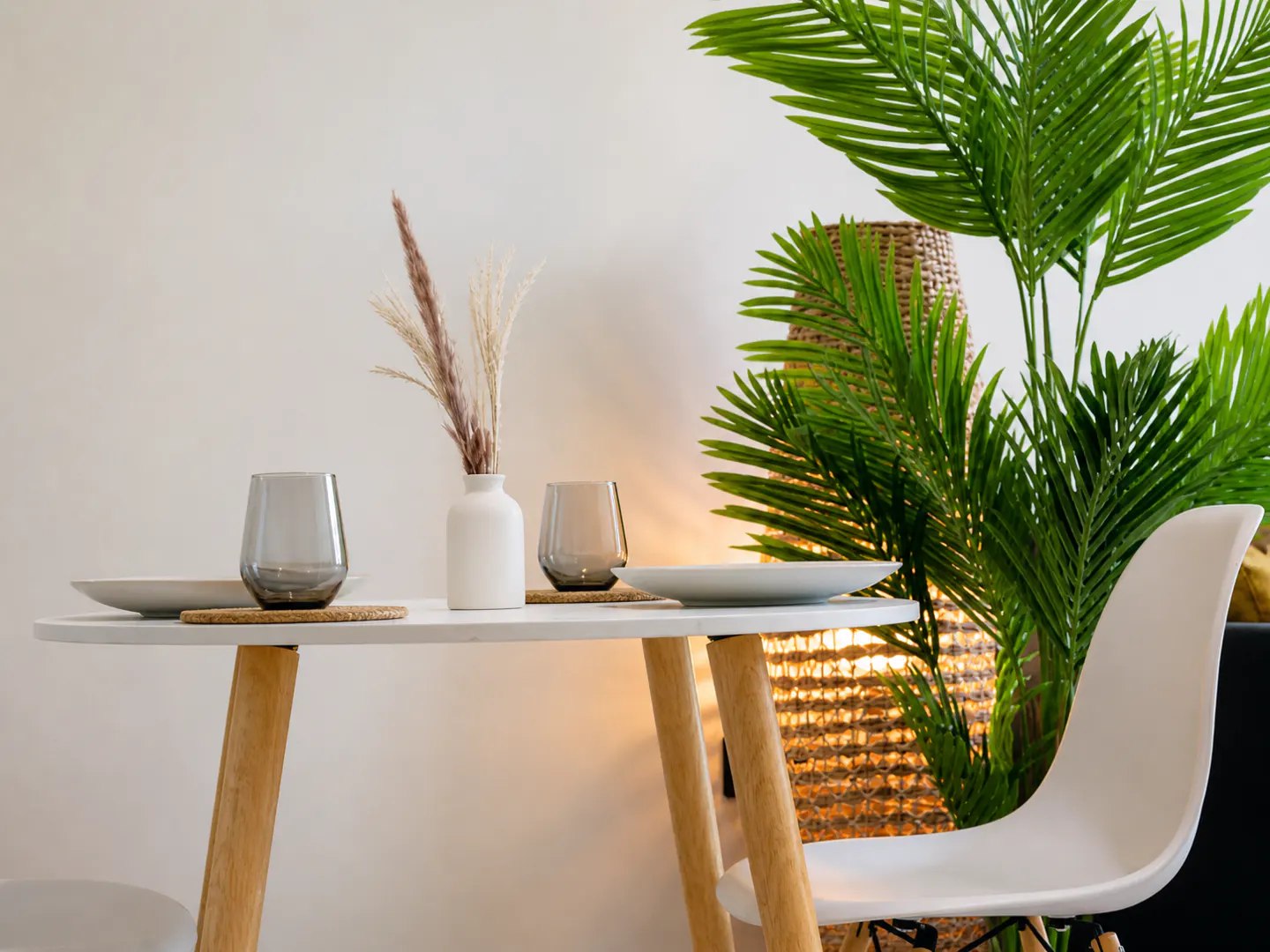 A modern dining area with a round white table, wooden legs, two gray glasses, plates, and a vase with dried flowers. A green palm plant adds a touch of nature.
