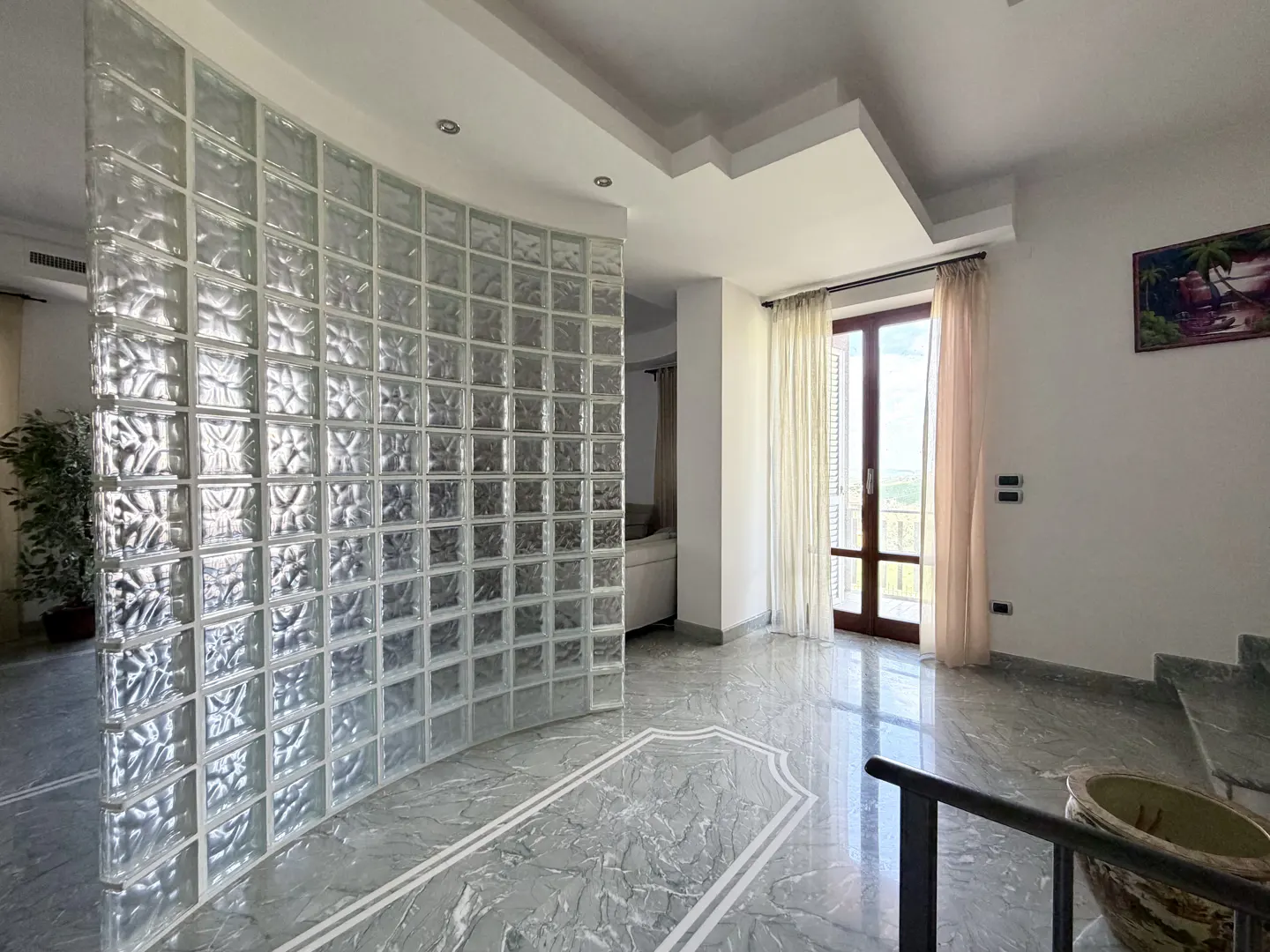 Bright foyer with a curved glass block wall, marble floors, and a balcony door with sheer curtains. Stairs lead up on the right.