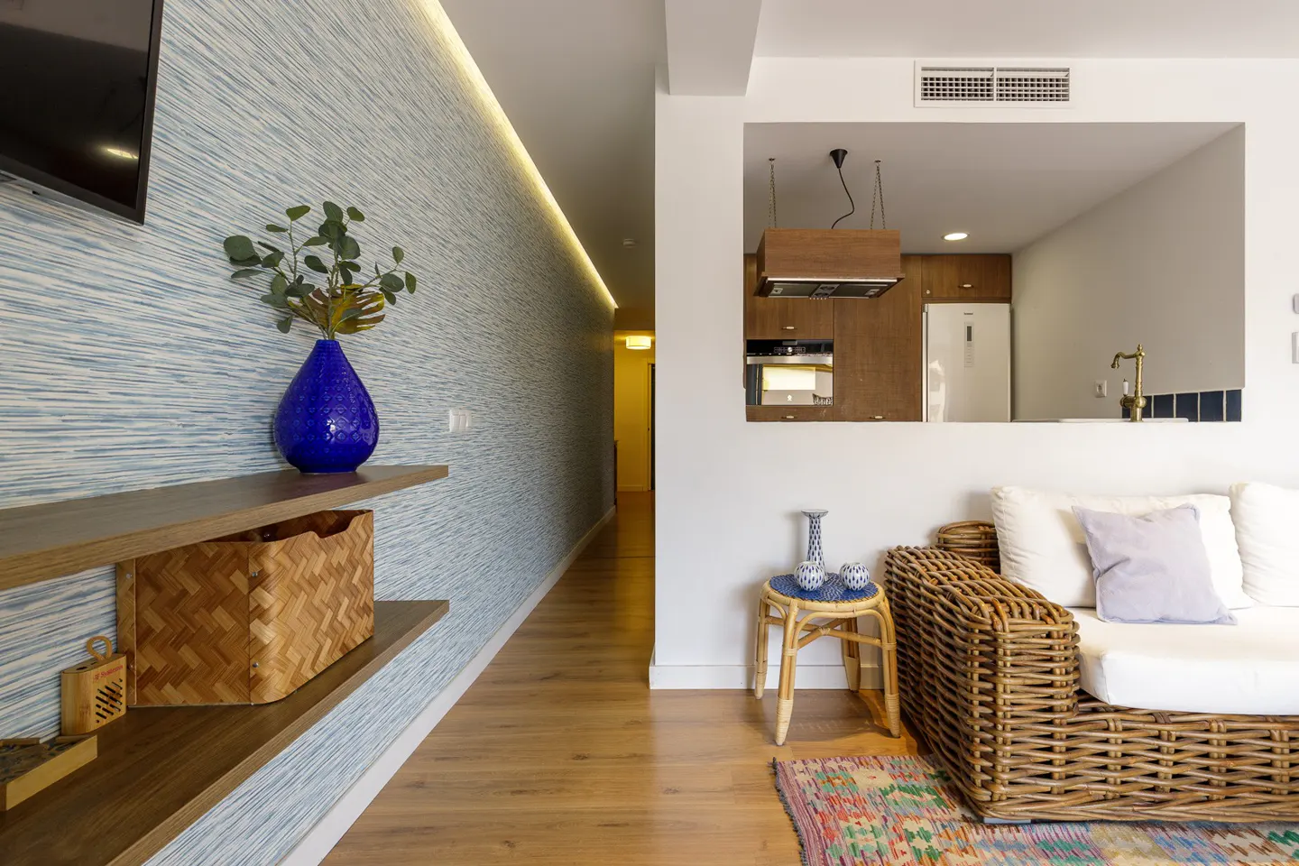 Living room with blue wallpaper, wood floors, and a wicker sofa. A blue vase sits on a shelf. The kitchen is visible through a cutout.