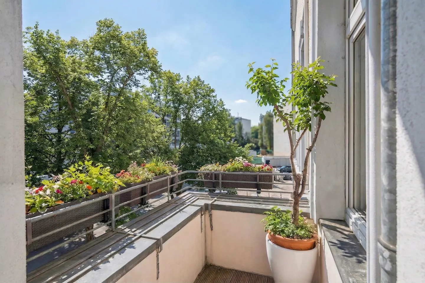 Balcony with flower boxes and a potted tree. Green trees and a blue sky are visible in the background.