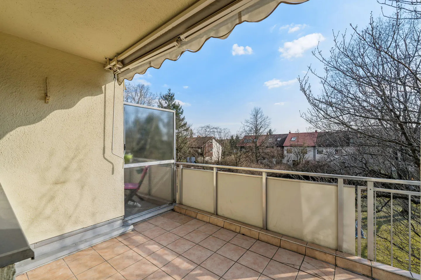 A balcony with beige tile flooring, a metal railing, and a retractable awning. Trees and houses are visible in the background.