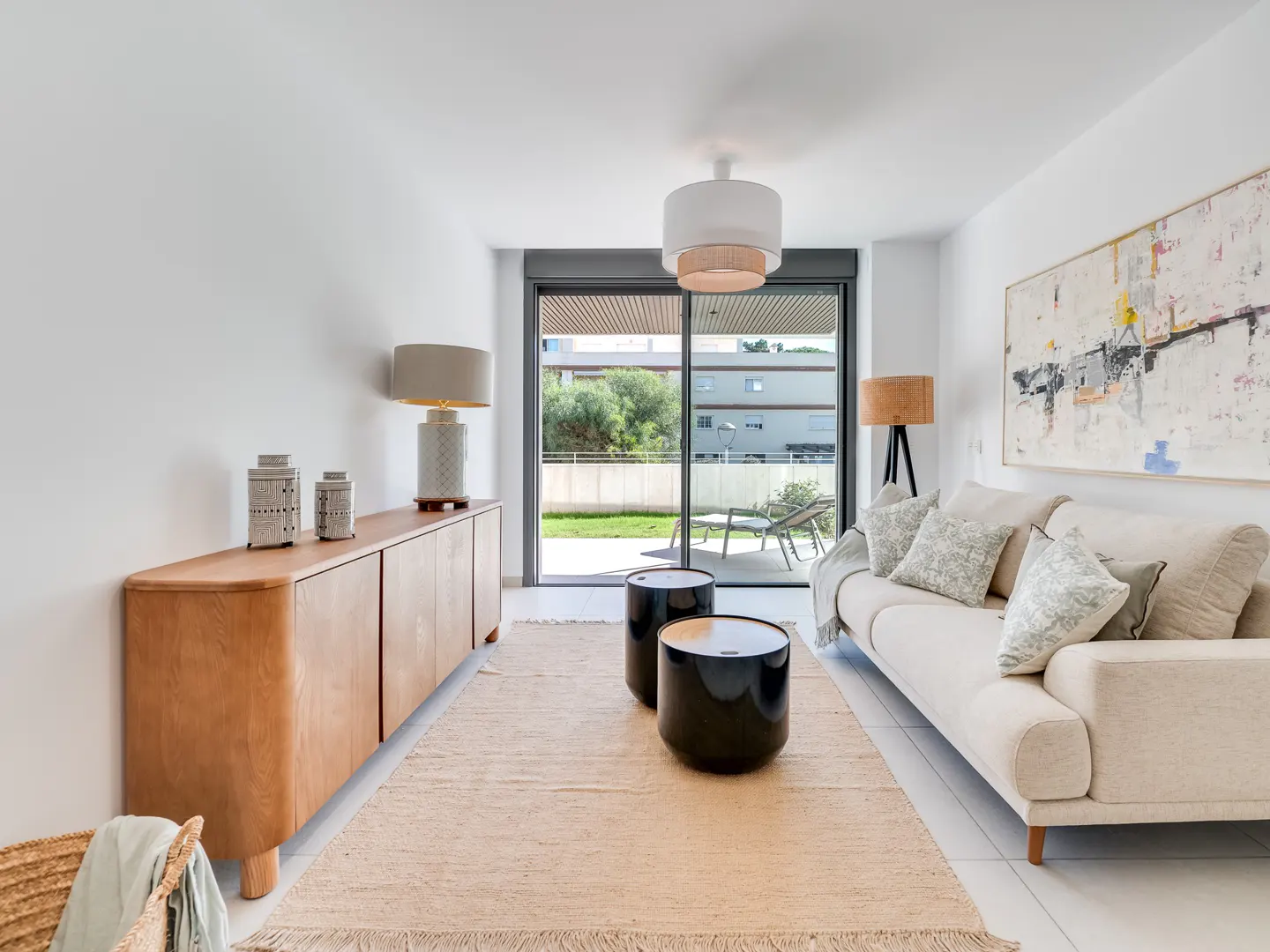 Bright living room with a beige sofa, black side tables, and a wooden cabinet. A large sliding glass door leads to a patio. Abstract art hangs on the white wall.