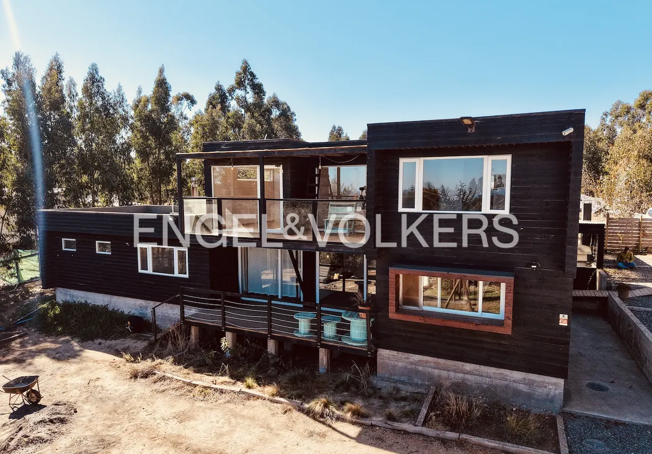 Two-story black house with white trim and large windows, surrounded by trees under a blue sky. "Engel & Volkers" logo overlay.