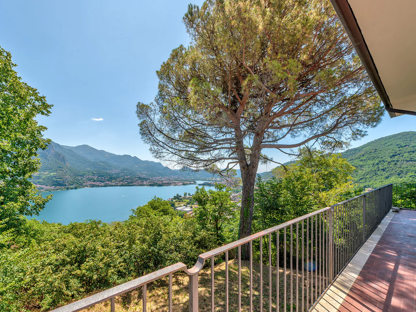 View from a balcony with a metal railing overlooking a lake, mountains, and trees on a sunny day.