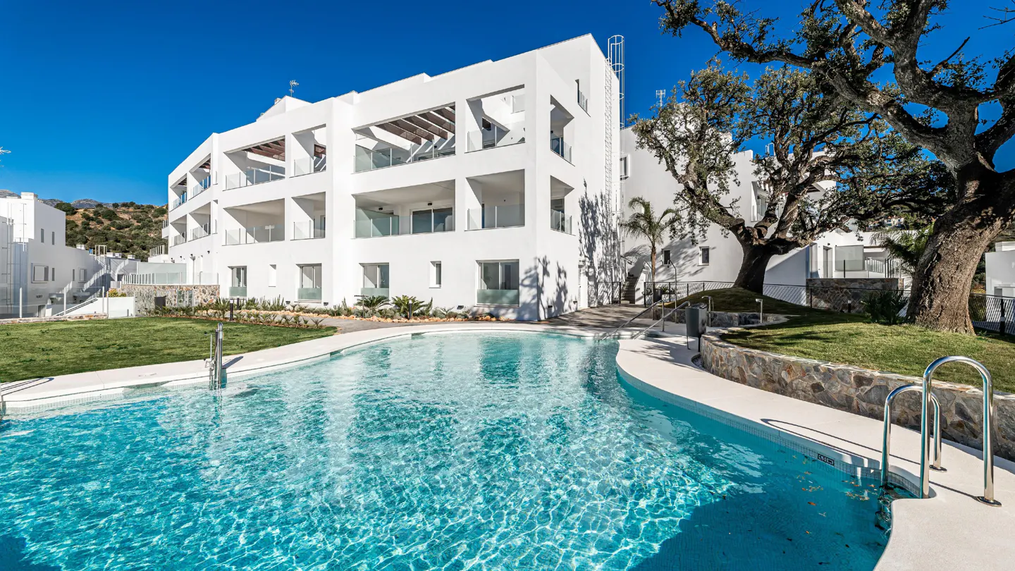 Modern white apartment building with balconies overlooks a turquoise pool and green lawn under a clear blue sky.