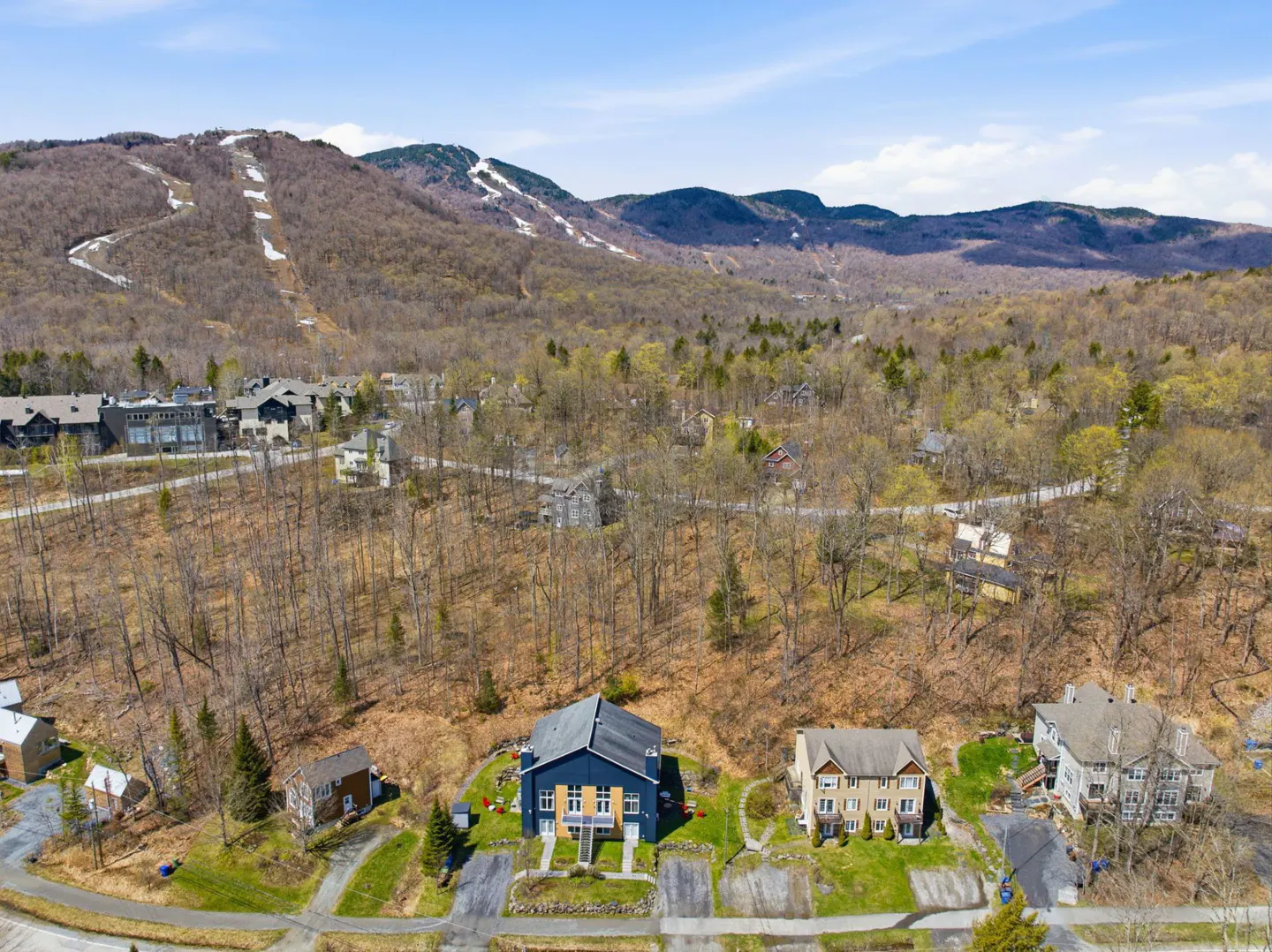Aerial view of houses near a mountain with ski trails, surrounded by trees under a blue sky.
