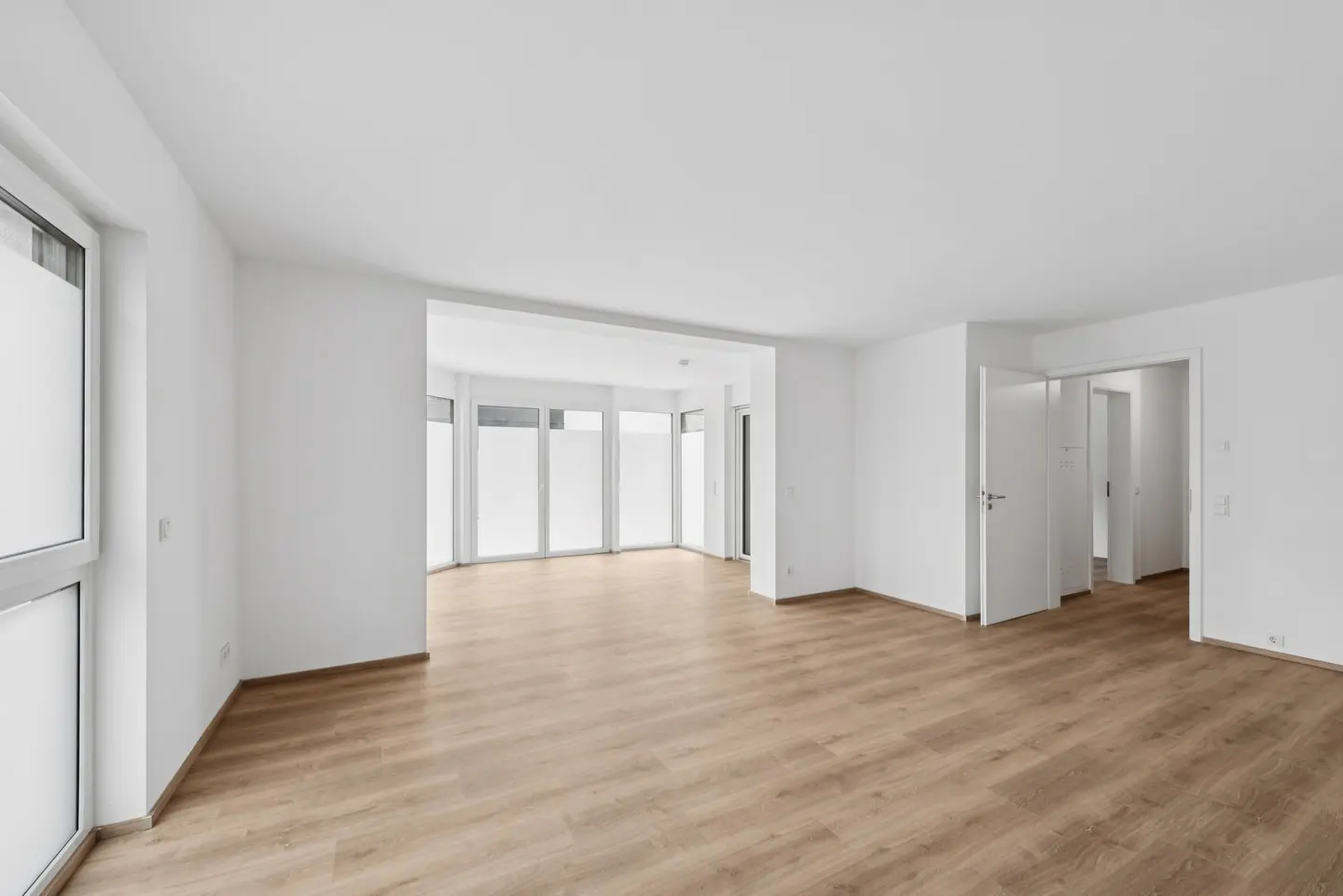 Bright, empty room with light wood floors, white walls, and large windows. Two open white doors lead to other rooms.