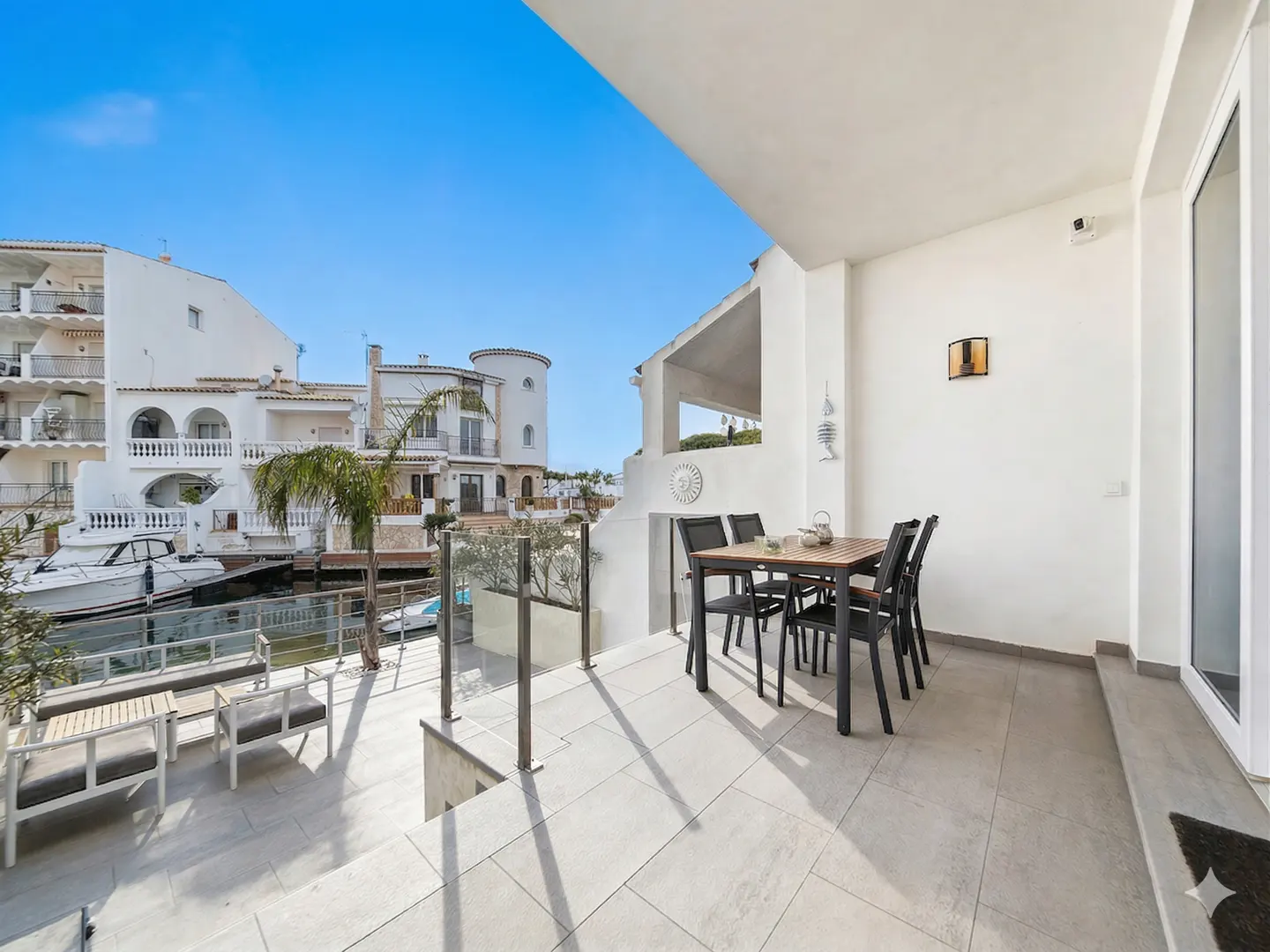Balcony view of a canal with boats, white buildings, and a blue sky. A table and chairs sit on the tiled balcony.