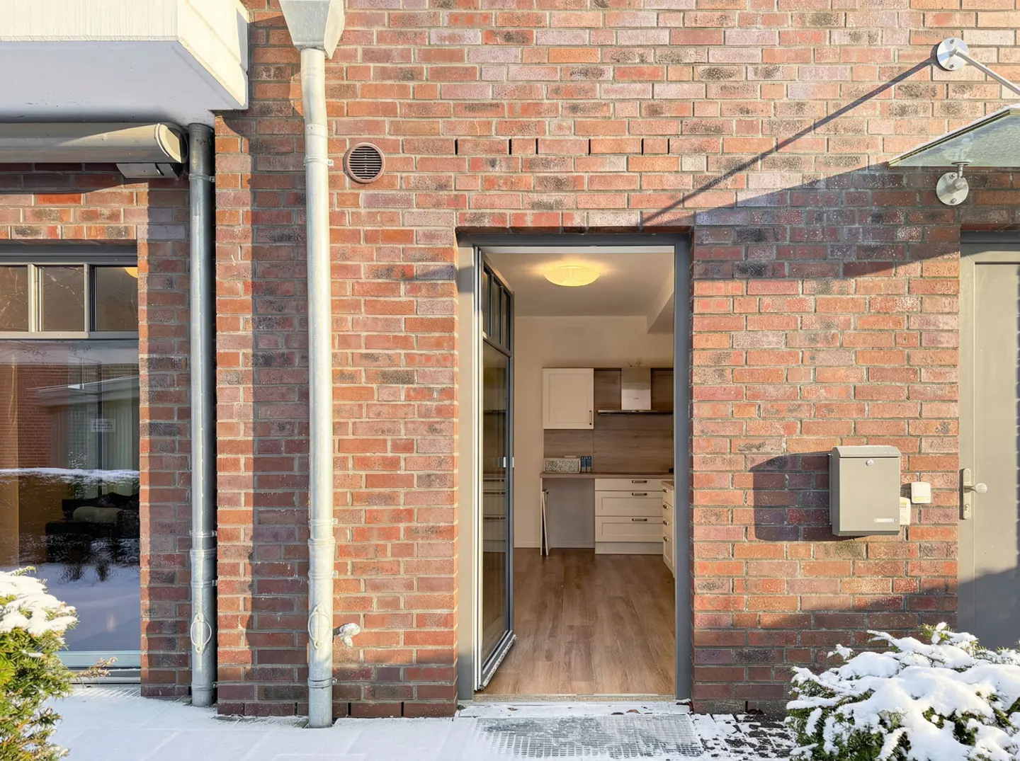 Exterior view of a brick house with an open door revealing a kitchen interior. Snow covers the ground.