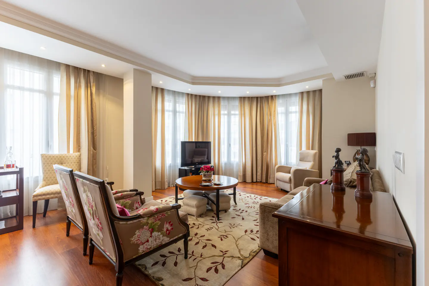 Bright living room with floral armchairs, a round wooden coffee table, beige curtains, and hardwood floors.