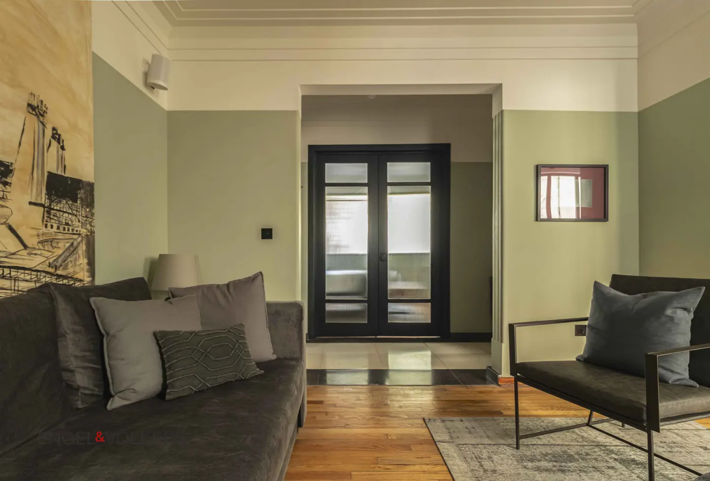 Living room with gray sofa, chair, and wood floors. A black framed double door is in the background. Walls are light green.