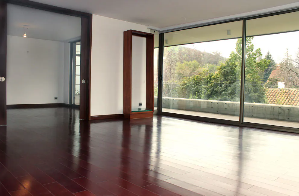 An empty room with dark wood floors, white walls, and a large window with a view of trees.