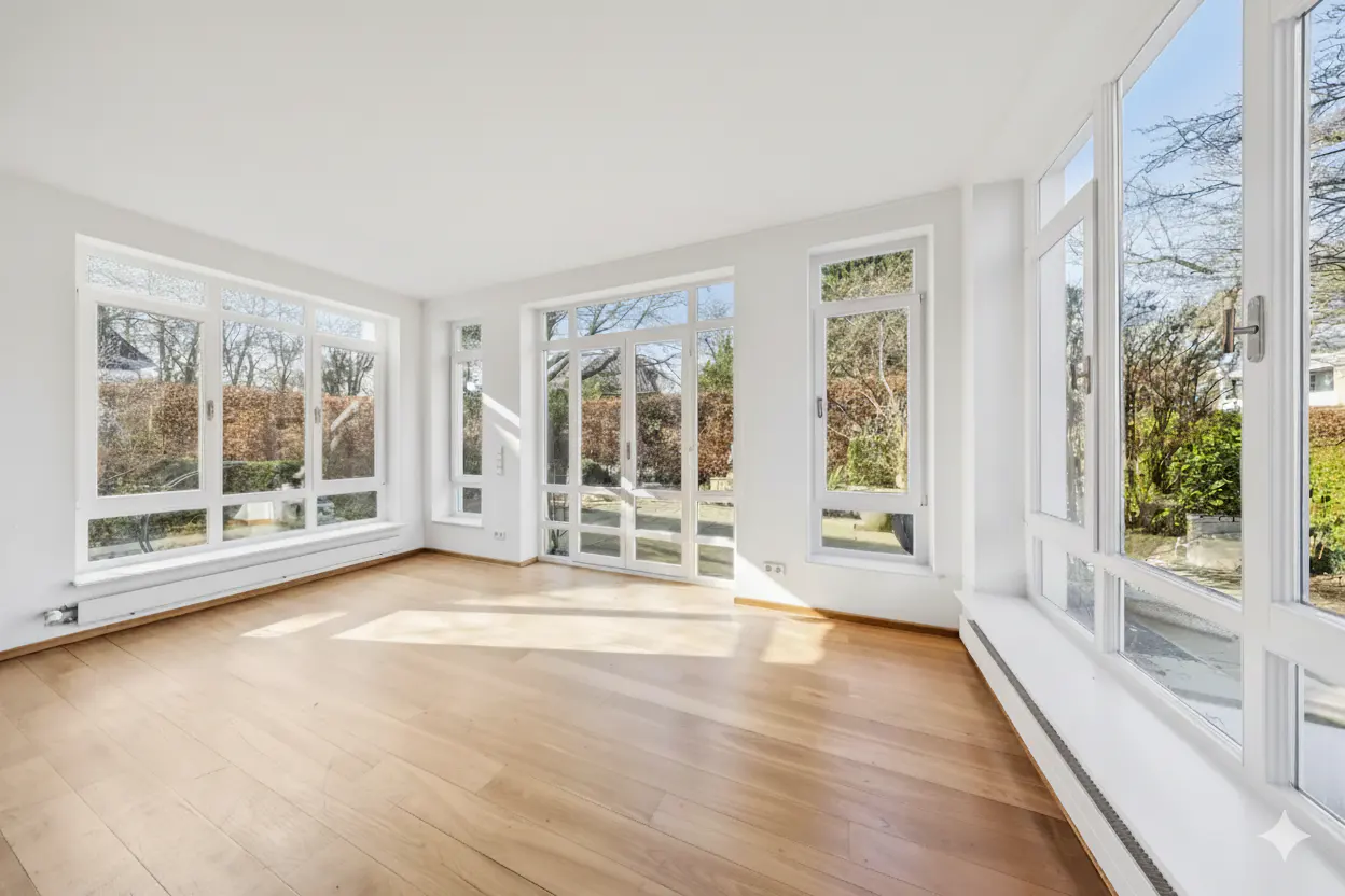 Bright, empty room with hardwood floors and white walls. Large windows offer views of a green, tree-filled yard. Sunlight streams in.