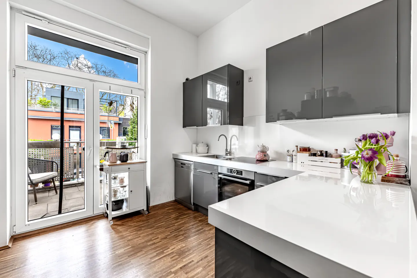 A modern kitchen with gray cabinets, white countertops, and wood floors. A balcony is visible through the open white doors.