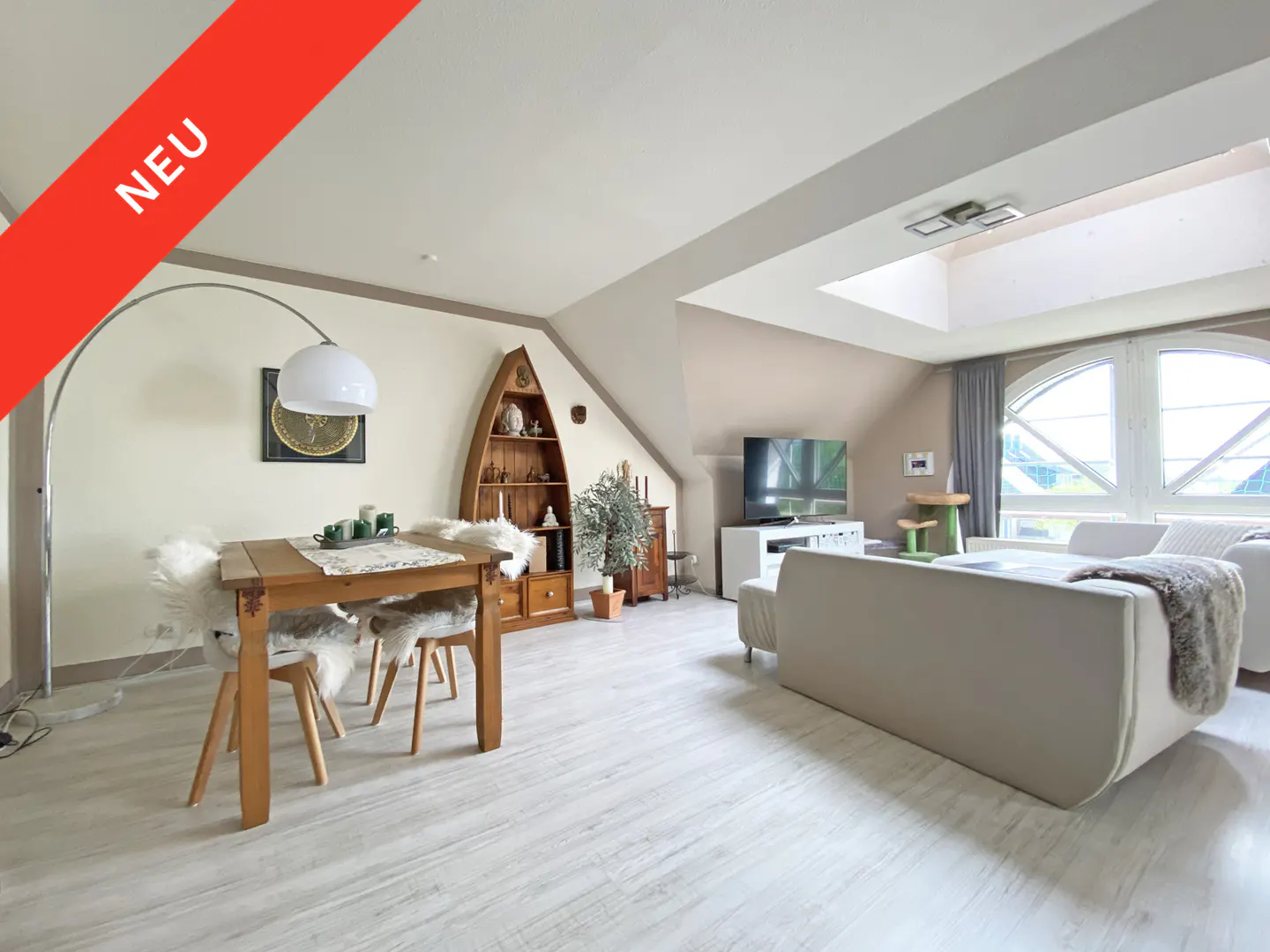 Bright living room with a wooden table, white chairs, a sofa, and a TV. A boat-shaped shelf adds a unique touch. "NEU" banner.
