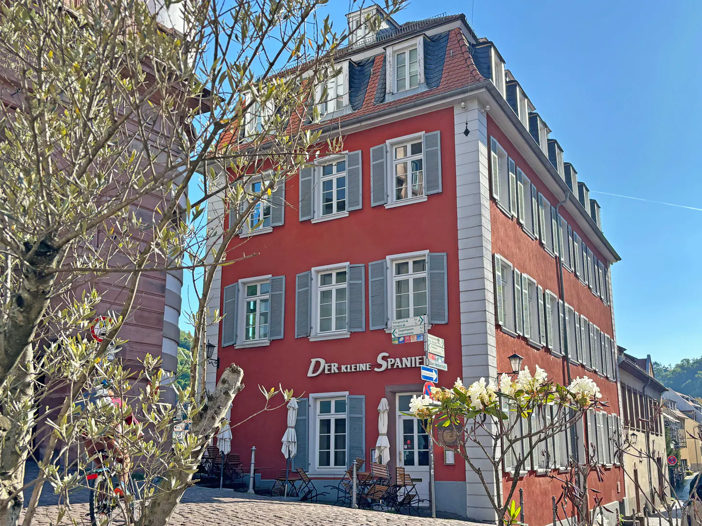 Exterior of "Der Kleine Spanier," a red building with grey shutters and a red tile roof, on a sunny day.