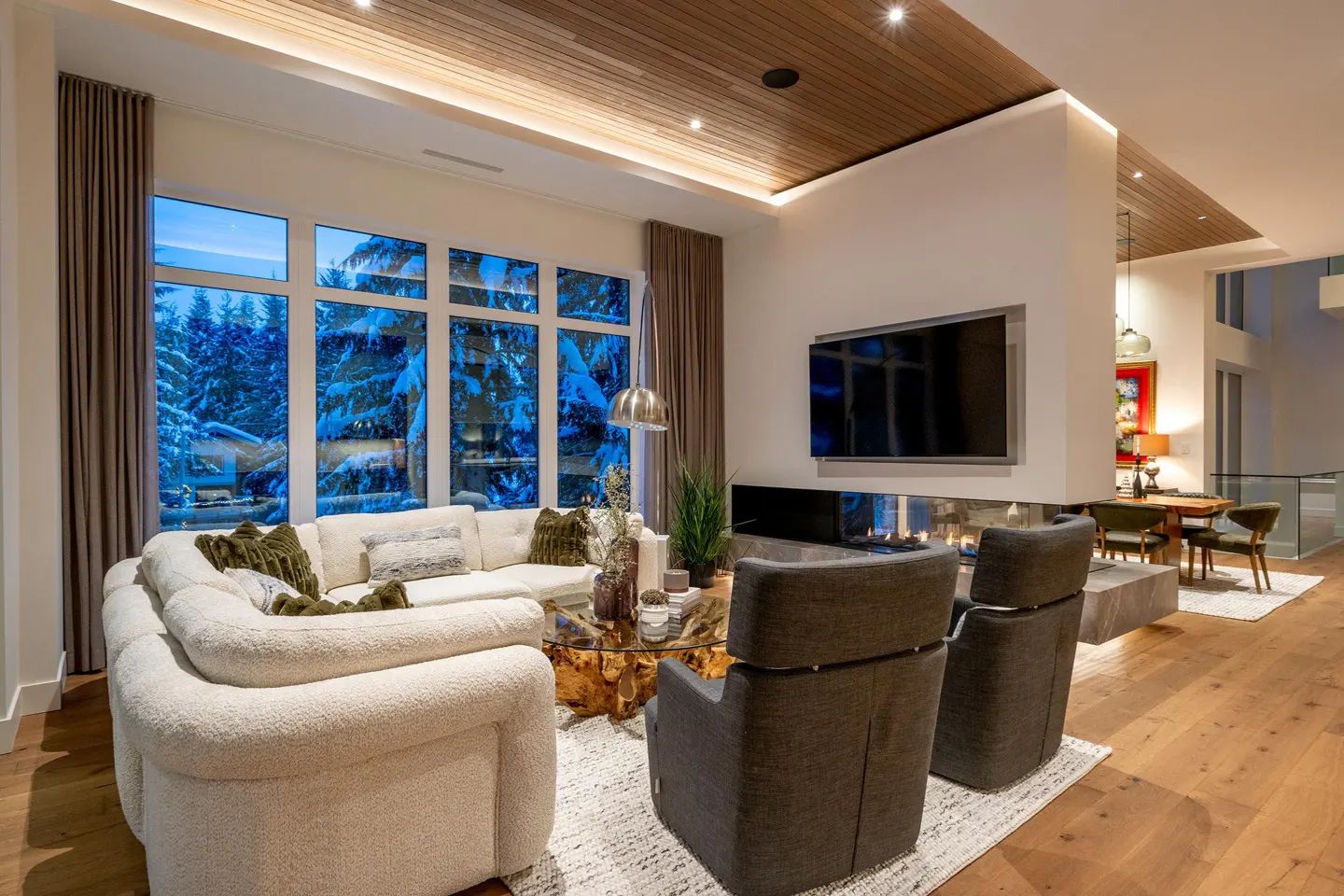 A modern living room with a white couch, gray chairs, and a fireplace. Large windows show snow-covered trees.