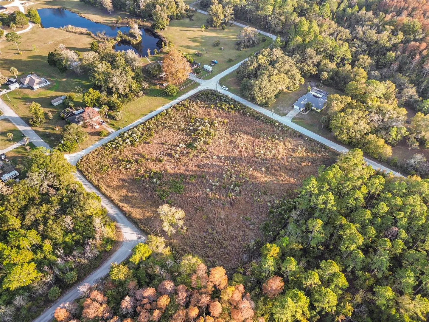 Aerial view of a large, rectangular vacant lot surrounded by trees, a small pond, and residential roads in a rural area.