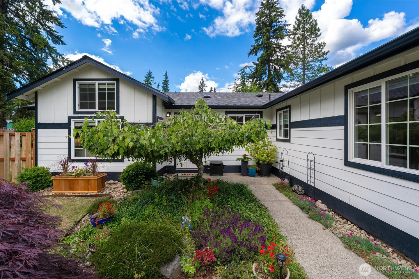 White house with dark trim, a concrete walkway, and a lush front garden with a small tree under a bright blue sky.