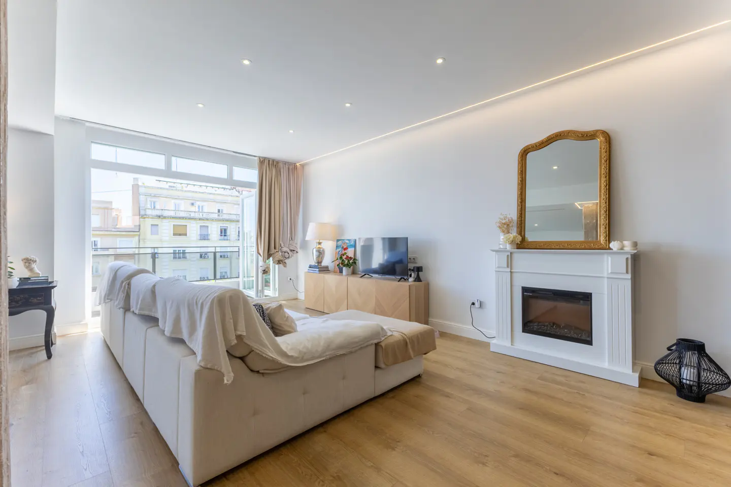 Bright living room with a white sofa, fireplace with gold-framed mirror, and a balcony overlooking city buildings.