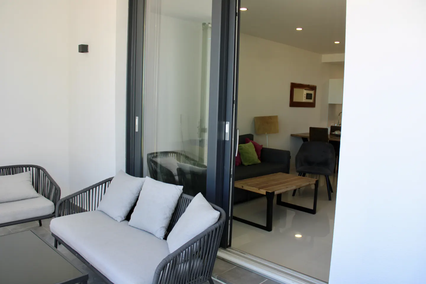 Balcony with gray wicker furniture and white cushions, sliding glass doors open to a living room with a gray sofa and wooden coffee table.