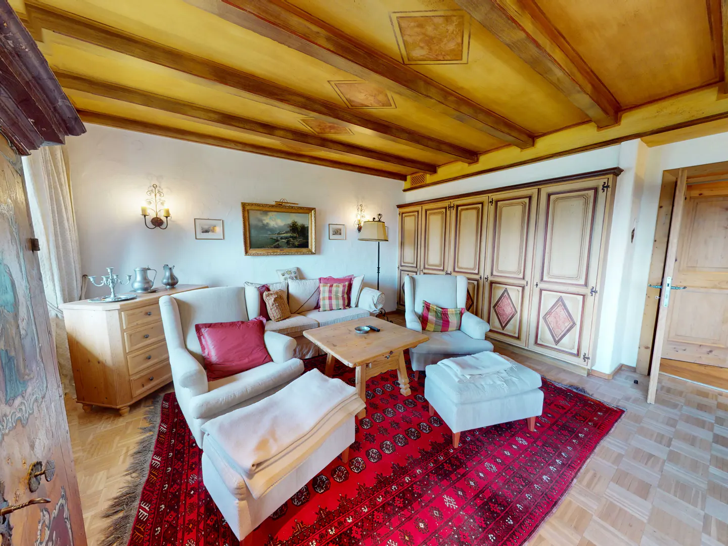 Living room with white armchairs, wooden coffee table, and red patterned rug under a yellow beamed ceiling.
