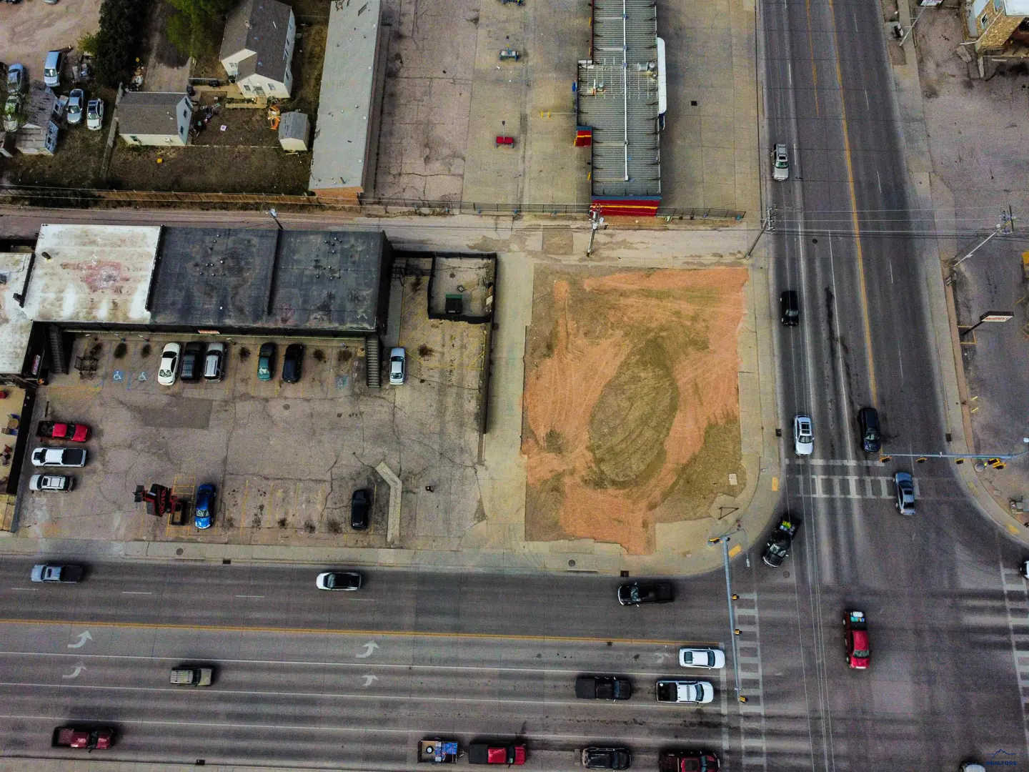 Aerial view of a vacant dirt lot next to a parking lot and commercial buildings near a busy multi-lane city street.