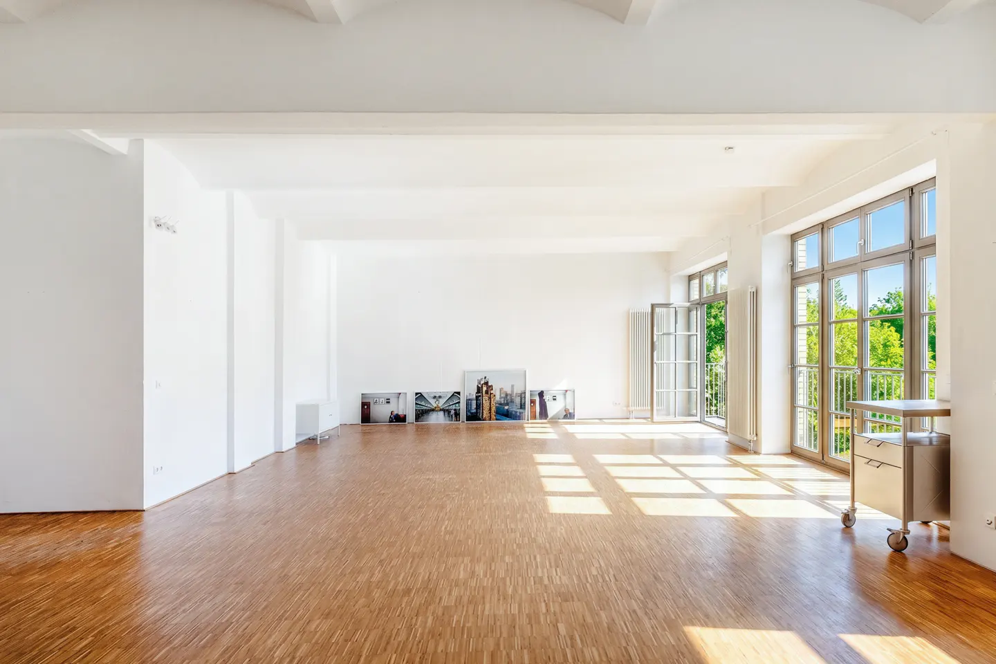 Bright, empty room with wood floors, white walls, and large windows. Sunlight streams in, illuminating the space. Pictures lean against the wall.