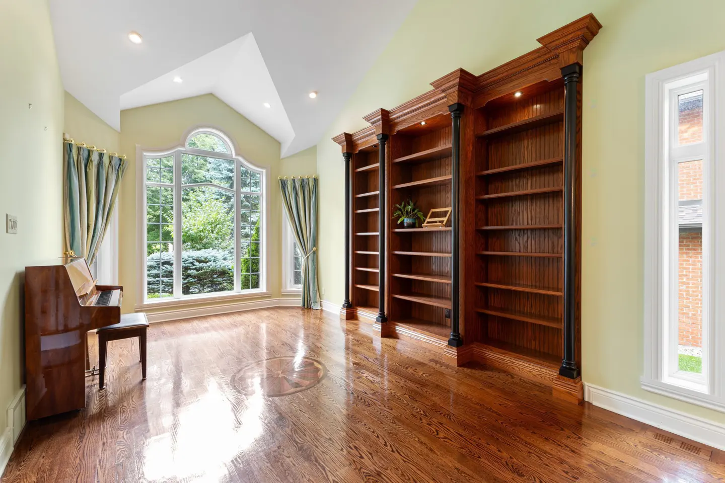 A bright room with hardwood floors, a piano, a large window with curtains, and three dark wood bookshelves.