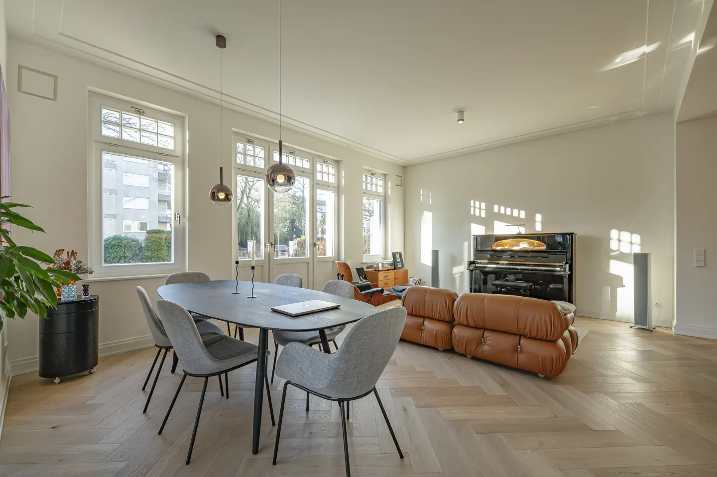 Bright, open-plan living and dining area with herringbone wood floors. A black dining table with gray chairs sits near large windows. A brown leather sofa and black piano are in the background.