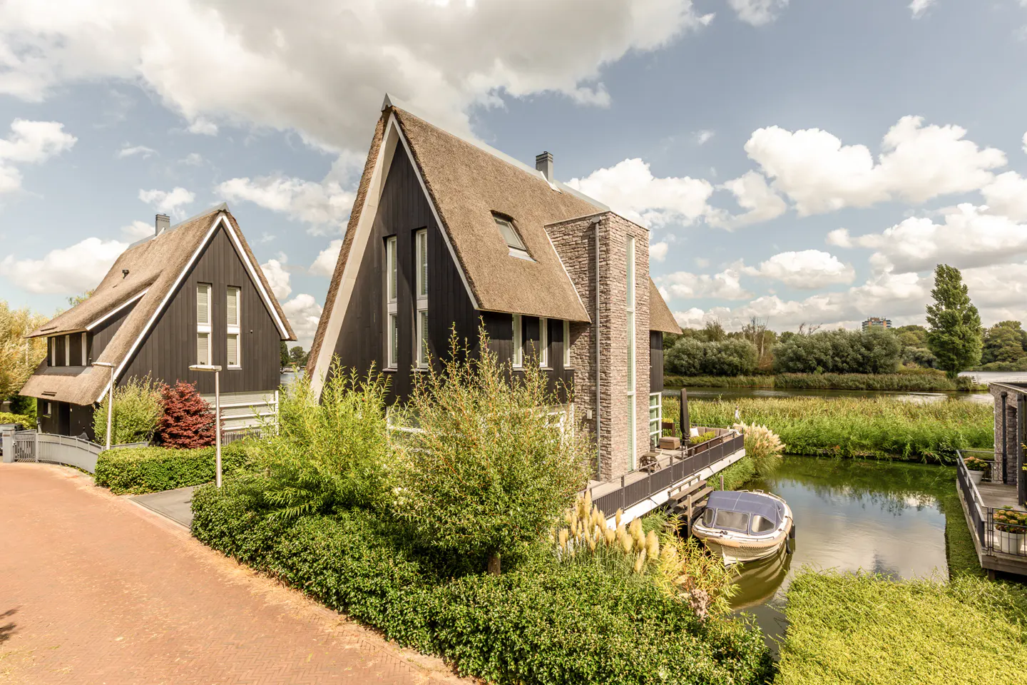 Two modern black houses with thatched roofs sit on a canal, with a boat docked nearby.