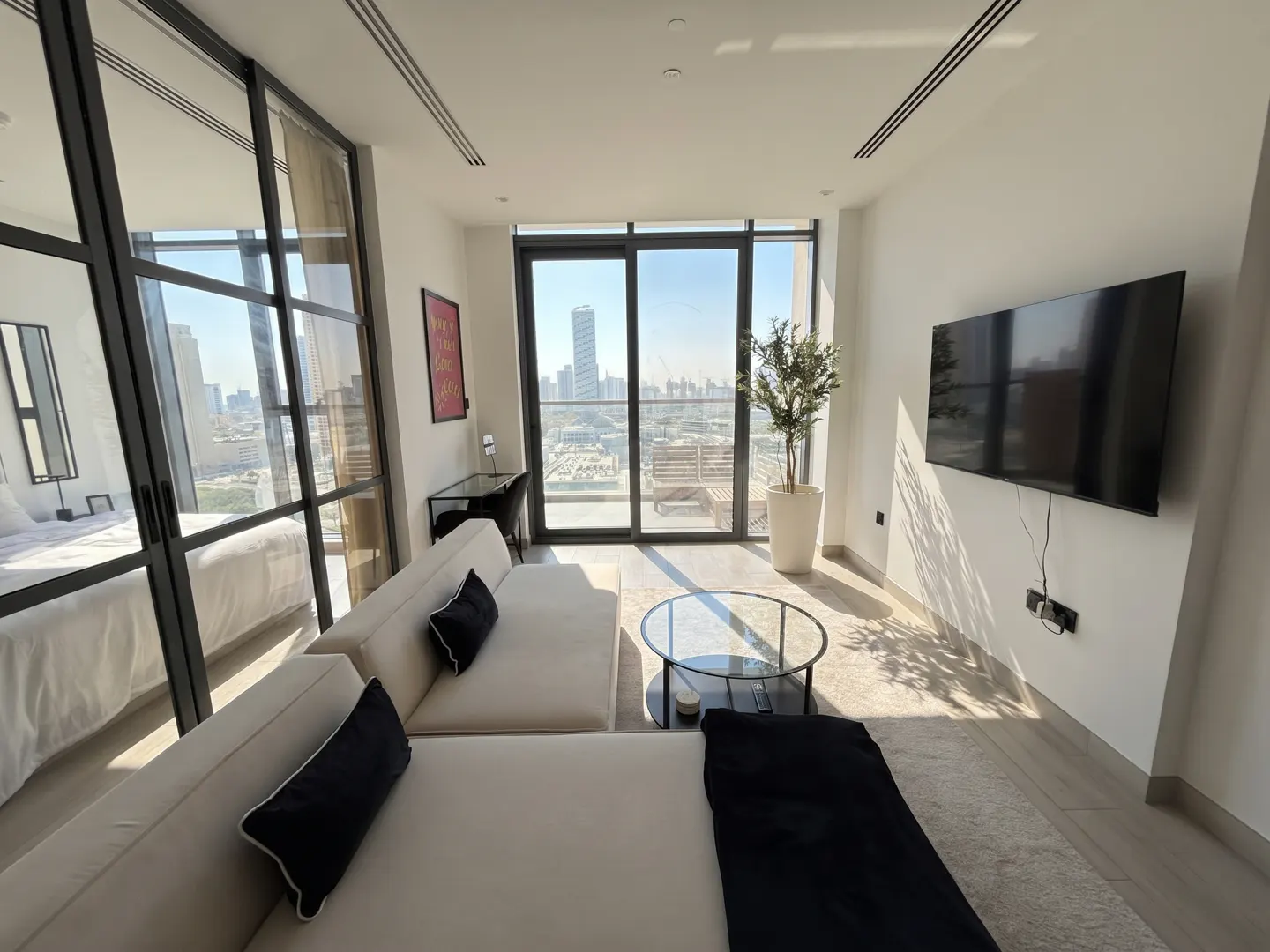 Bright, modern apartment living room with a white sofa, black pillows, and a city view through large windows. A TV hangs on the wall.