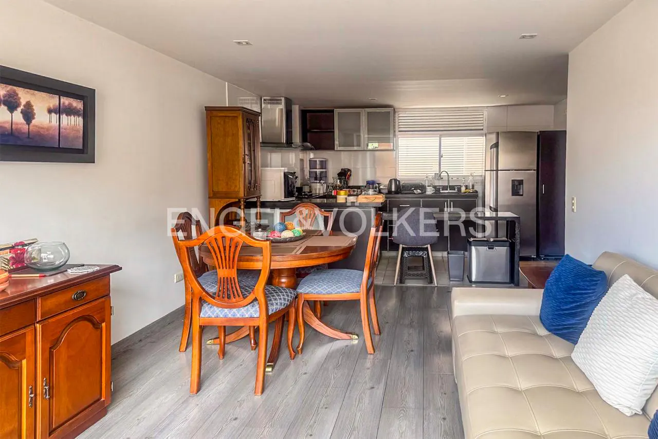 Open-plan living area with a wooden dining set, a beige sofa with blue pillows, and a modern kitchen in the background.