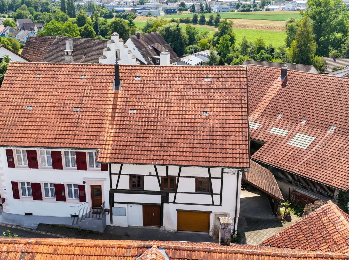 Aerial view of a white half-timbered house with a red tile roof, adjacent to a barn in a rural village setting.