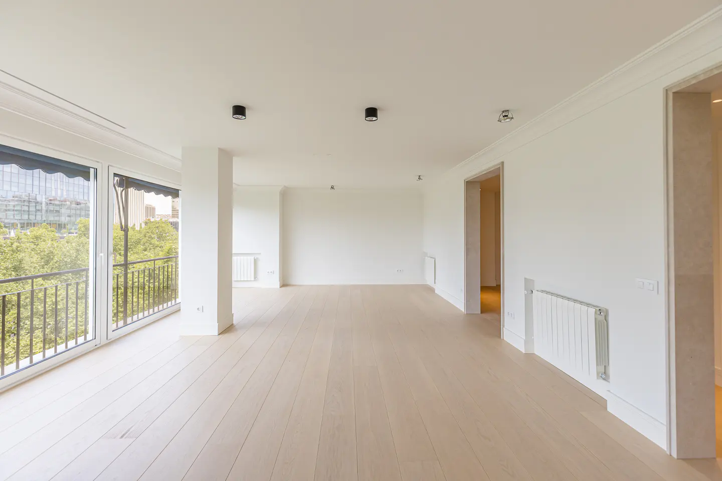 Bright, empty room with light wood floors, white walls, and a balcony with a city view. Black ceiling lights and white radiators are visible.