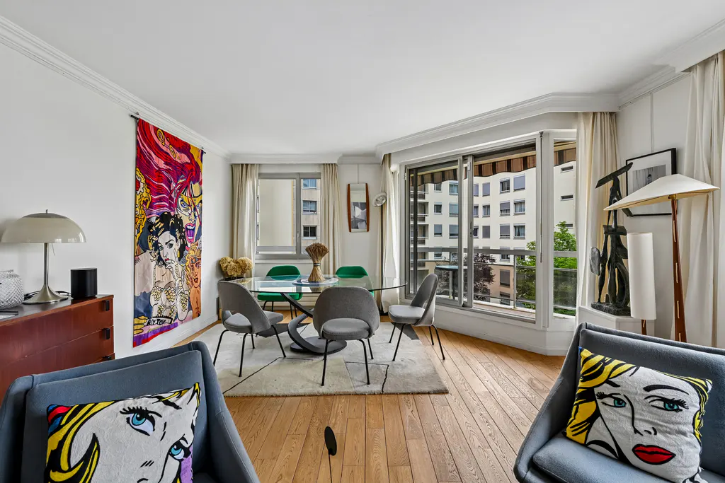 Bright living room with wood floors, a glass dining table, gray chairs, and a colorful pop art tapestry on the wall. Large windows overlook a city view.