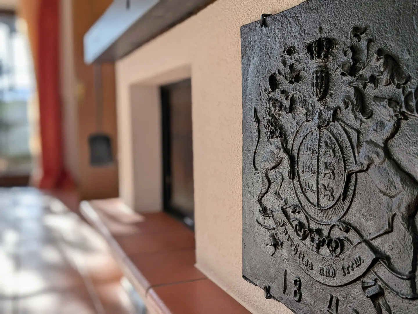 Close-up of a black, ornate coat of arms plaque on a beige wall next to a fireplace with a red-tiled hearth.