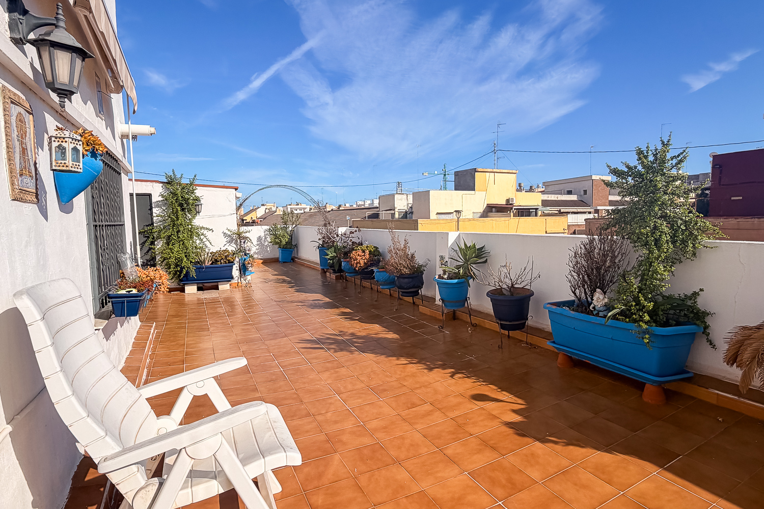 Sunny rooftop terrace with terracotta tiles, a white lounge chair, and a row of blue planters filled with various plants. [AI-generated text]