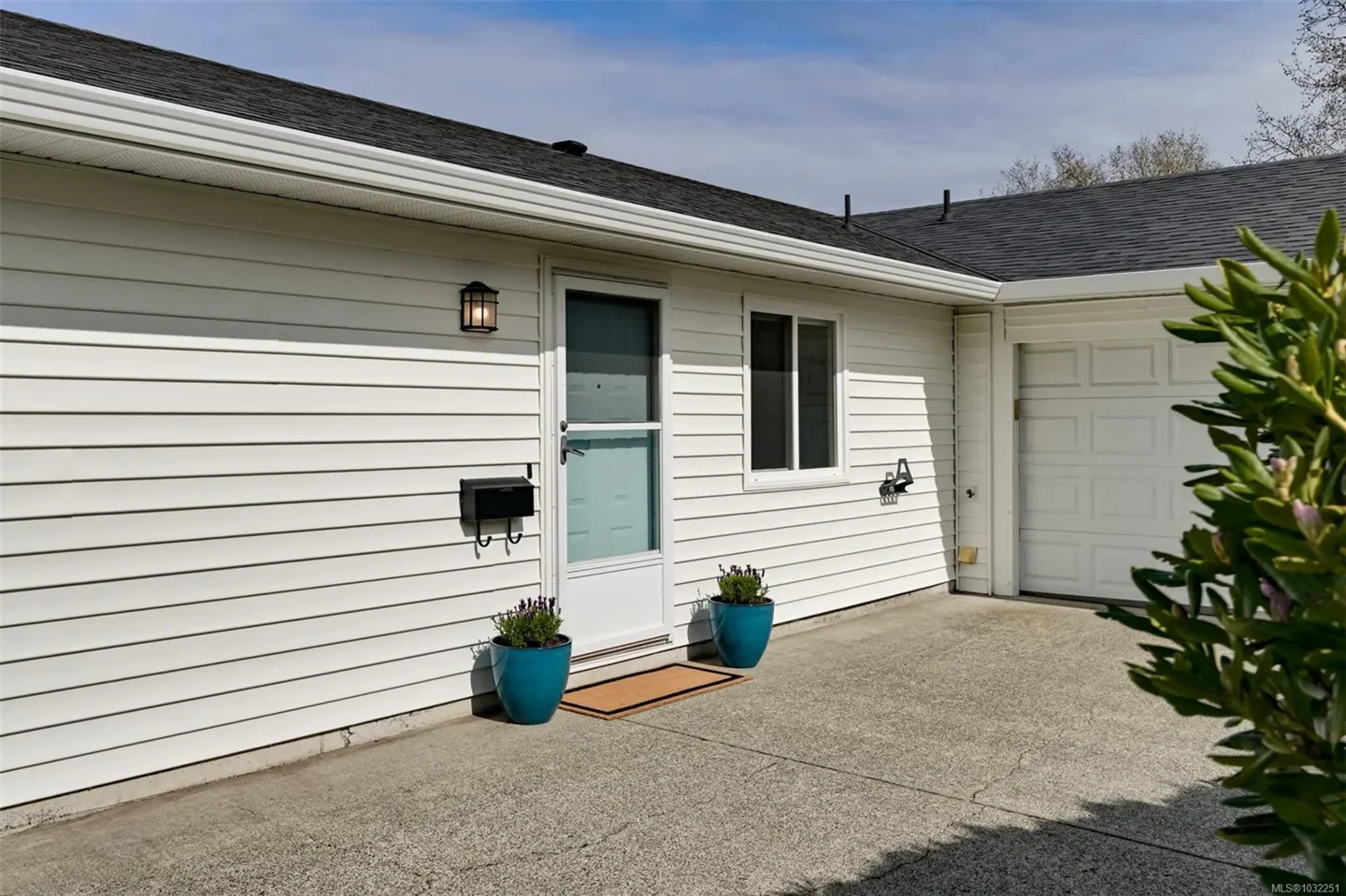 Exterior view of a white house with a black roof, a white garage door, and a light blue front door. Two blue pots with lavender flank the door.
