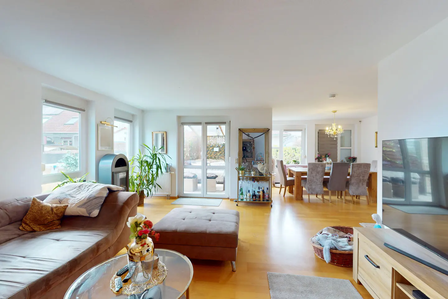 Bright, open-concept living room with hardwood floors, brown sofa, ottoman, and dining area with table and chairs. White walls and natural light.