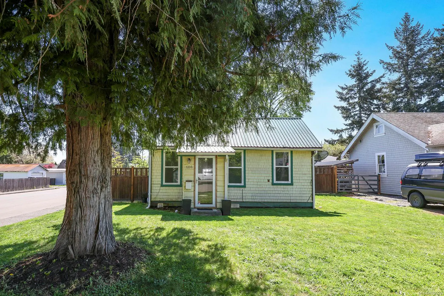 A small, light yellow house with green trim and a metal roof sits on a green lawn, shaded by a large tree.