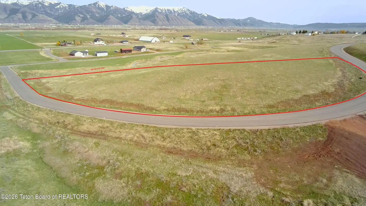 Aerial view of a grassy lot outlined in red, with a curved road, houses, and snow-capped mountains in the background.