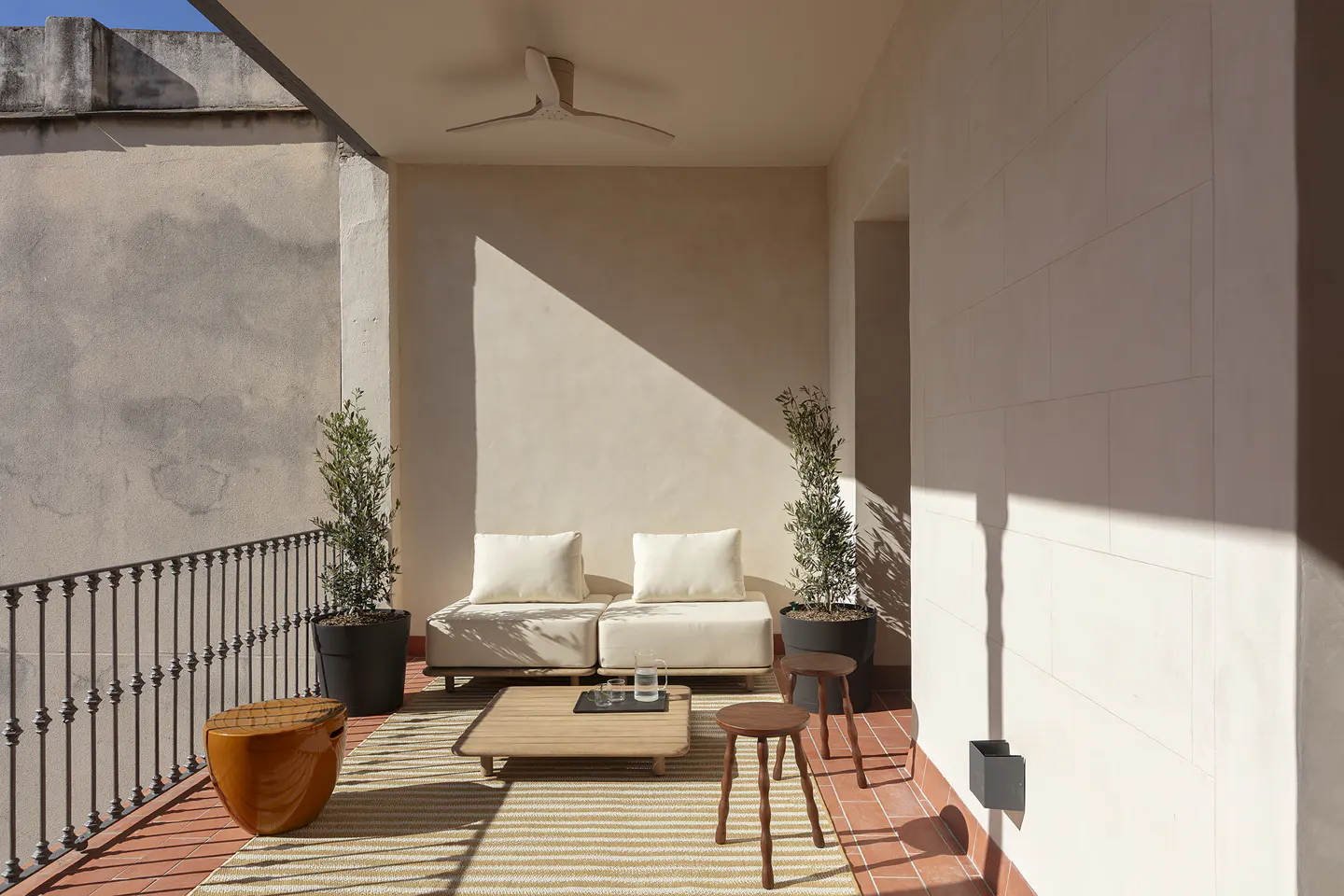 A sunlit balcony with two cream sofas, a wooden table, and potted olive trees. A striped rug covers the red tile floor.