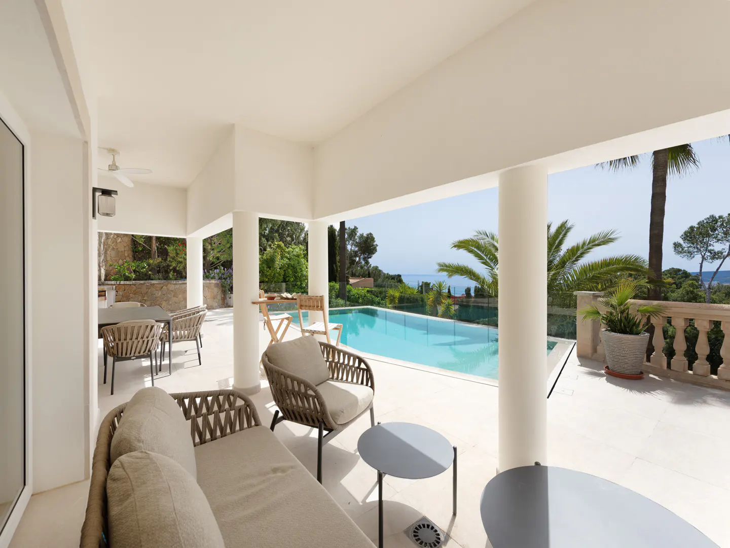 Covered patio with wicker furniture overlooks a blue pool and lush greenery. White pillars support the roof.