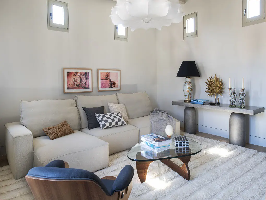 Living room with a white sectional sofa, glass coffee table, and blue chair on a white rug. A gray console table with a lamp and decor is against the wall.