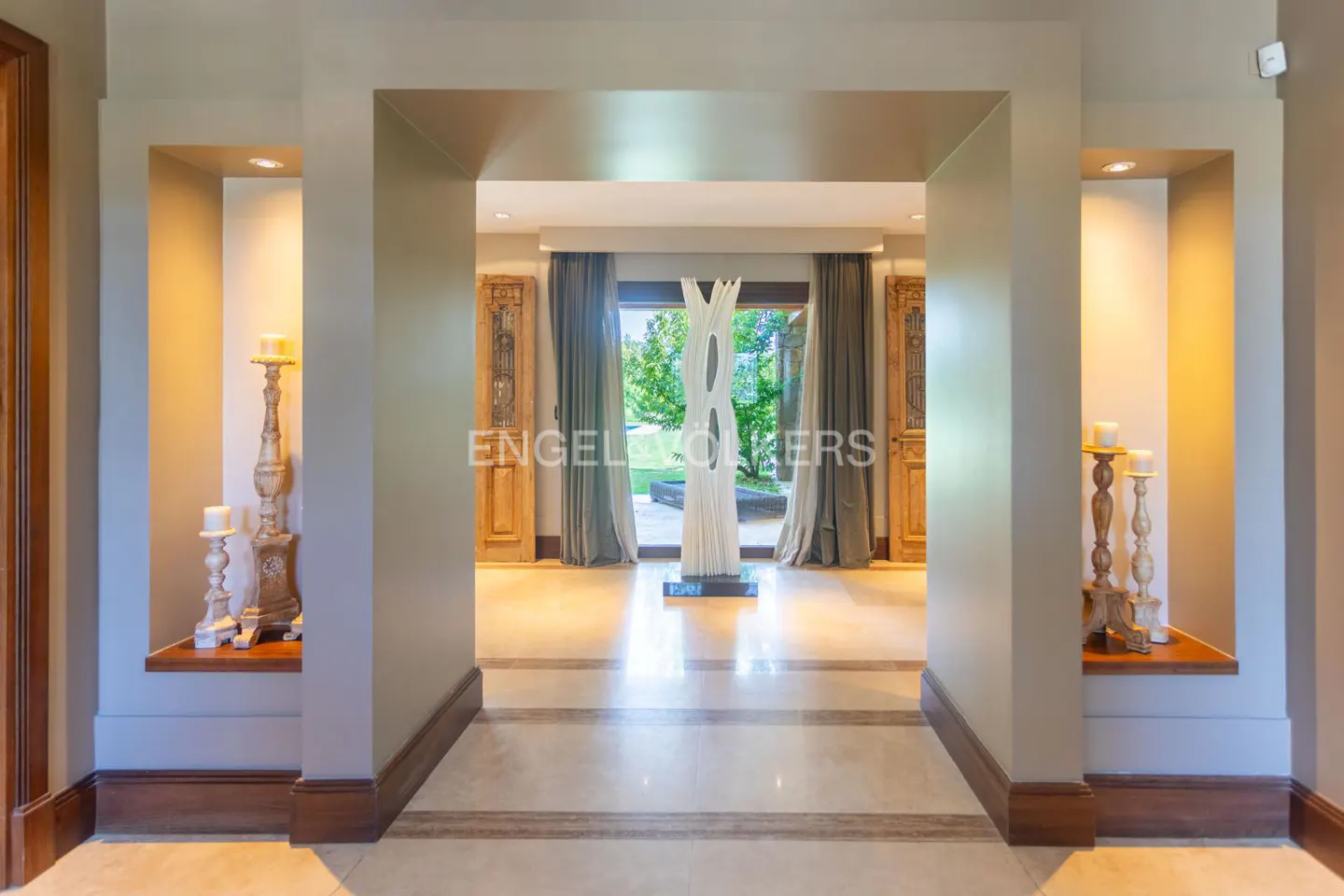 Foyer view with marble floors, beige walls, and recessed lighting. A white sculpture stands in front of open doors leading to a green garden.