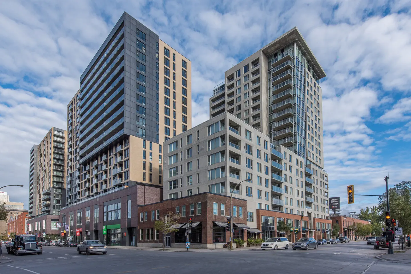 Cityscape view of modern high-rise apartment buildings with balconies, shops below, and cars on the street.