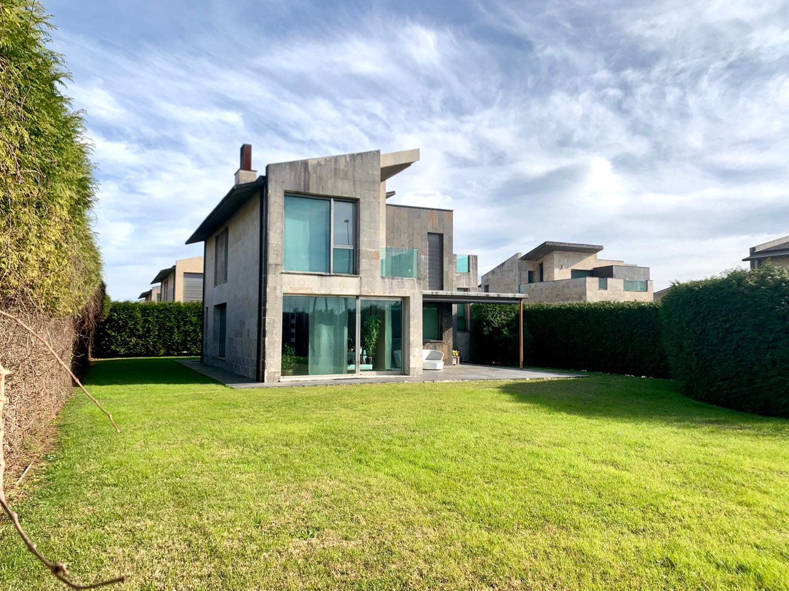 Modern two-story concrete house with large glass windows, a patio, and a spacious green lawn under a cloudy blue sky.
