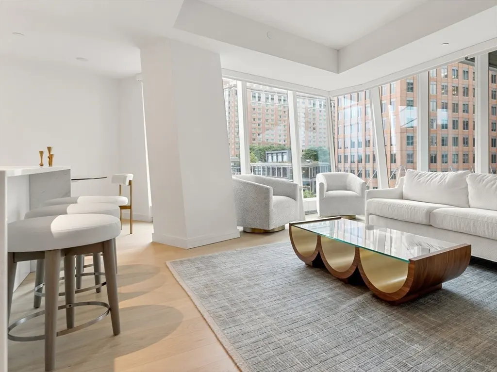 Bright, modern living room with white sofa, chairs, and a unique coffee table on a gray rug. Large windows offer a city view.